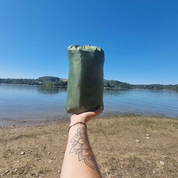 Une personne tenant un tapis de camping vert devant un paysage de lac, mettant en avant un équipement de plein air portable et pratique pour les voyages.